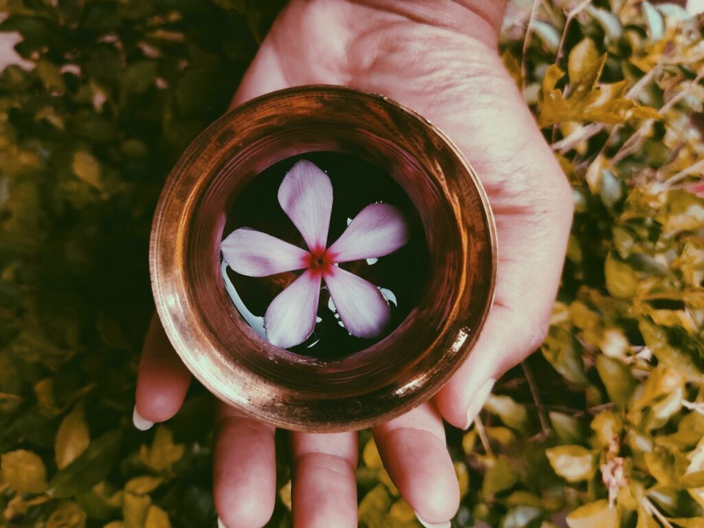 A close-up of a hand holding a brass bowl with a purple flower floating inside, surrounded by greenery.
